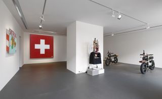 Wide view of 'The Pack' installation by Tom Sachs featuring two electric motorcycles, a hot drinks machine with hair and a dress, wall art of the red and white Swiss flag and another piece of wall art with a colourful map and red text in a room with grey flooring and white walls