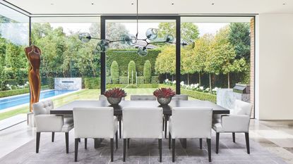 A minimalist dining room with a large brown table, white chairs, two succulents on the table, a statement light fixture and gold sculpture, with large floor to ceiling windows and a garden with a manicured lawn with a swimming pool seen behind