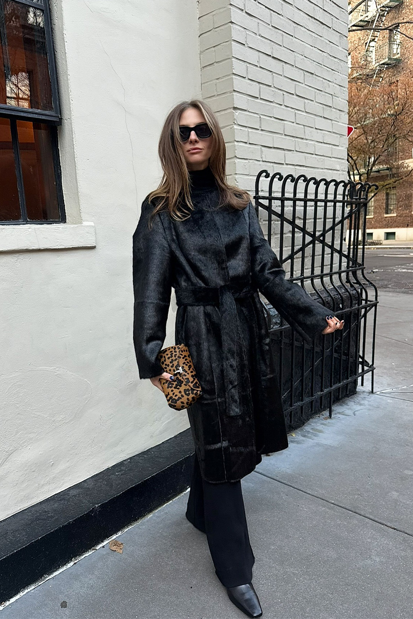 West Village winter trends are shown in a street style photo of a woman standing in front of a white building in her winter outfit, wearing a black pony hair coat layered over a black turtleneck and black wide-leg trousers styled with black pointed toe ankle boots, black sunglasses, and a cheetah print calf-hair clutch bag
