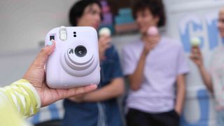 Fujifilm Instax Mini 12 in the hand with blurred subjects in background