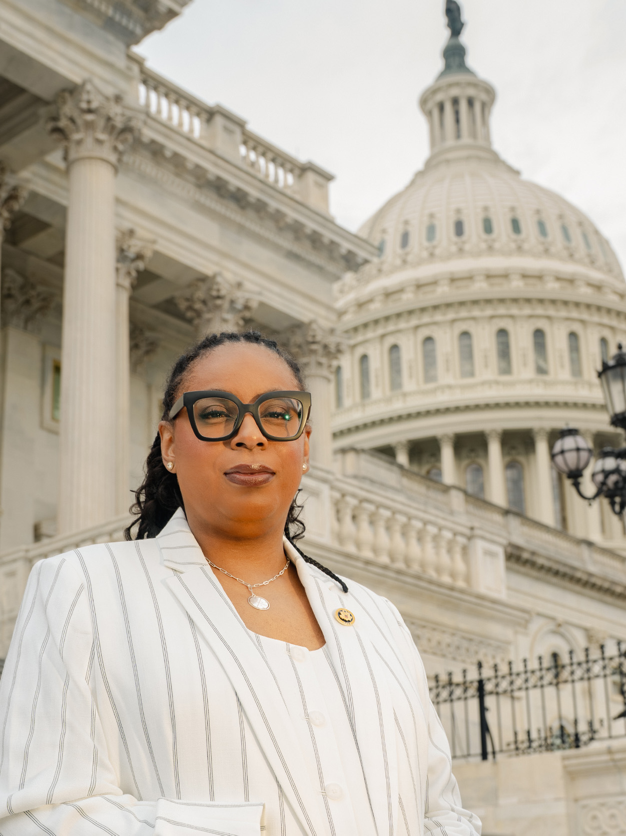 A close-up portrait of LaMonica McIver outdoors. She wears a white pinstripe blazer, dark-framed glasses, and a congressional pin. The Capitol dome is behind her against an overcast sky.