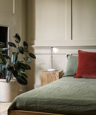 A bedroom with beige wall panels, a green potted plant, a white table lamp, and sage green bedding with a crushed velvet red throw pillow on it