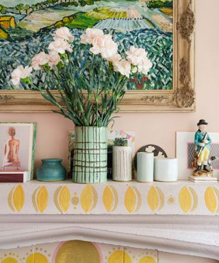 A light pink and yellow patterned mantlepiece with a countryside painted print above it and a green checked vase of light pink roses, three candles, and photo frames on it