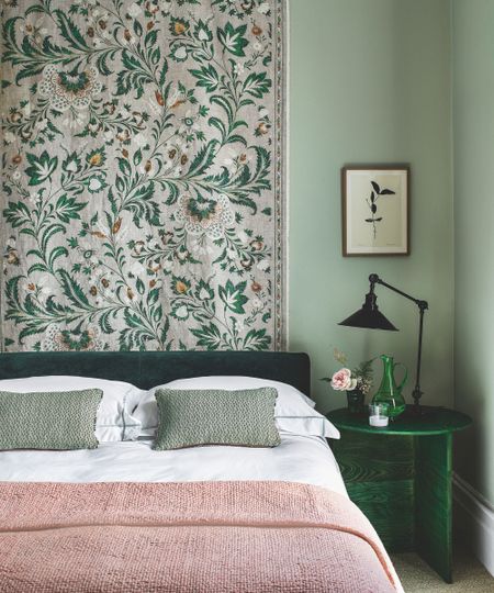 A green bedroom with a green velvet bed with white bedding, a pink blanket and green cushions, a patterned hanging tapestry behind the bed, a green bedside table with a green glass jug, pink flowers and a black lamp, with a small botanical artwork above. The walls are color drenched sage green.