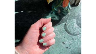 A close-up of Digital Beauty Editor, Fiona McKim's hand with short sage-green nails, pictured in front of a green marbled table