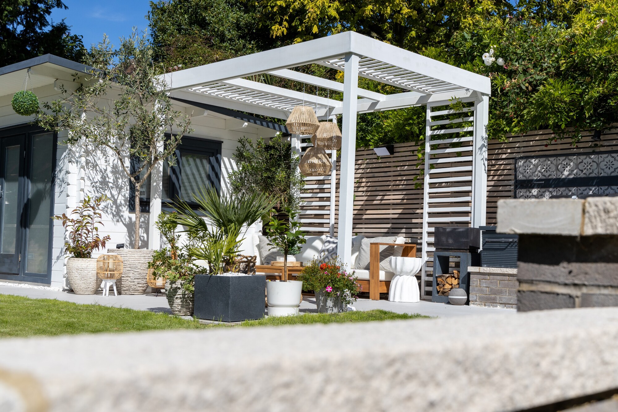 A modern garden with a white outhouse building and a white pergola attached to offer shade to an outdoor seating area