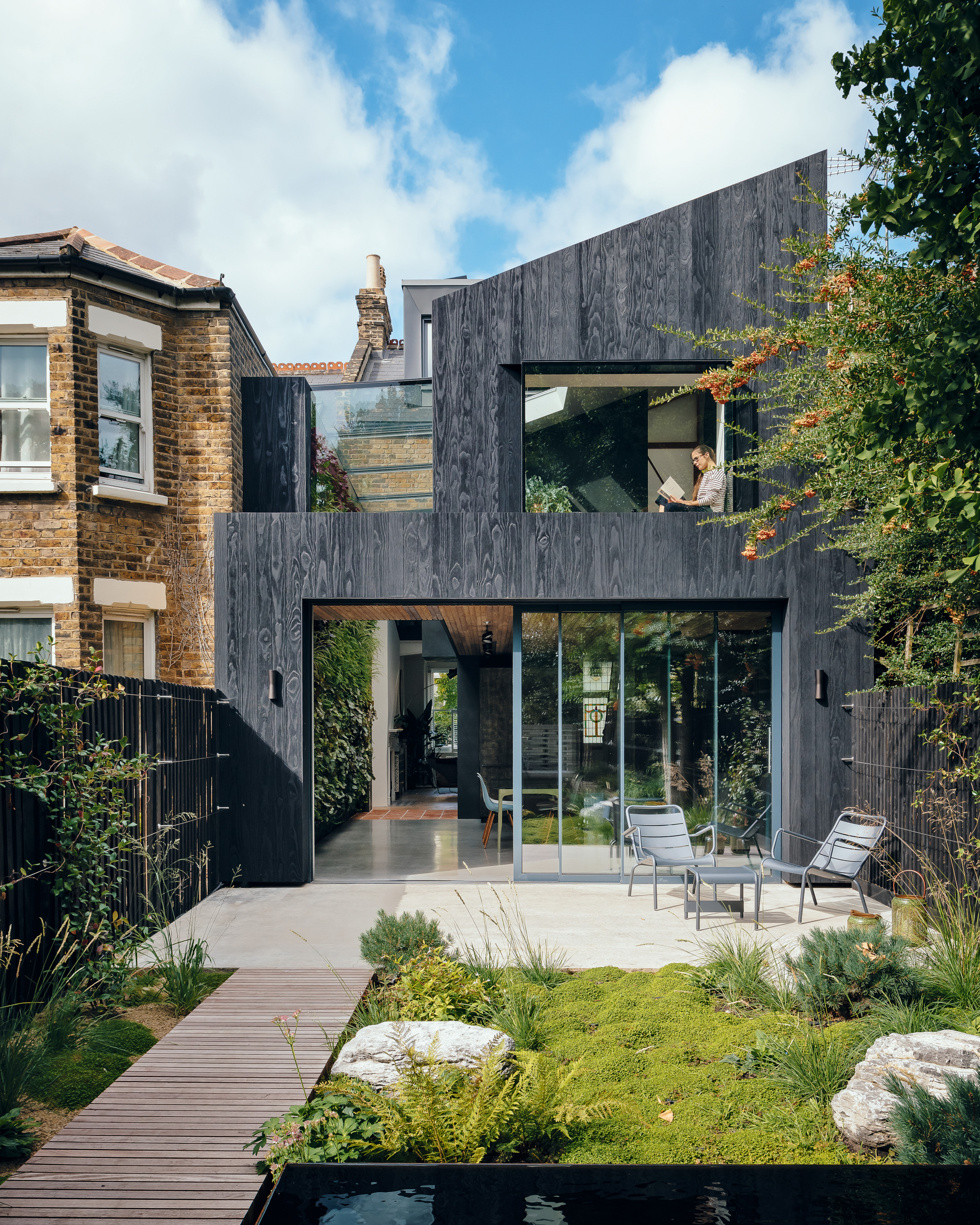 Charred wood-clad two-story extension leading to patio area with pale stone tiles, mossy grass area and slatted wood path