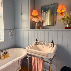 Half-panelled bathroom with traditional pedestal sink, roll top bath, shelf with vases of flowers, wall mirror and two wall lights