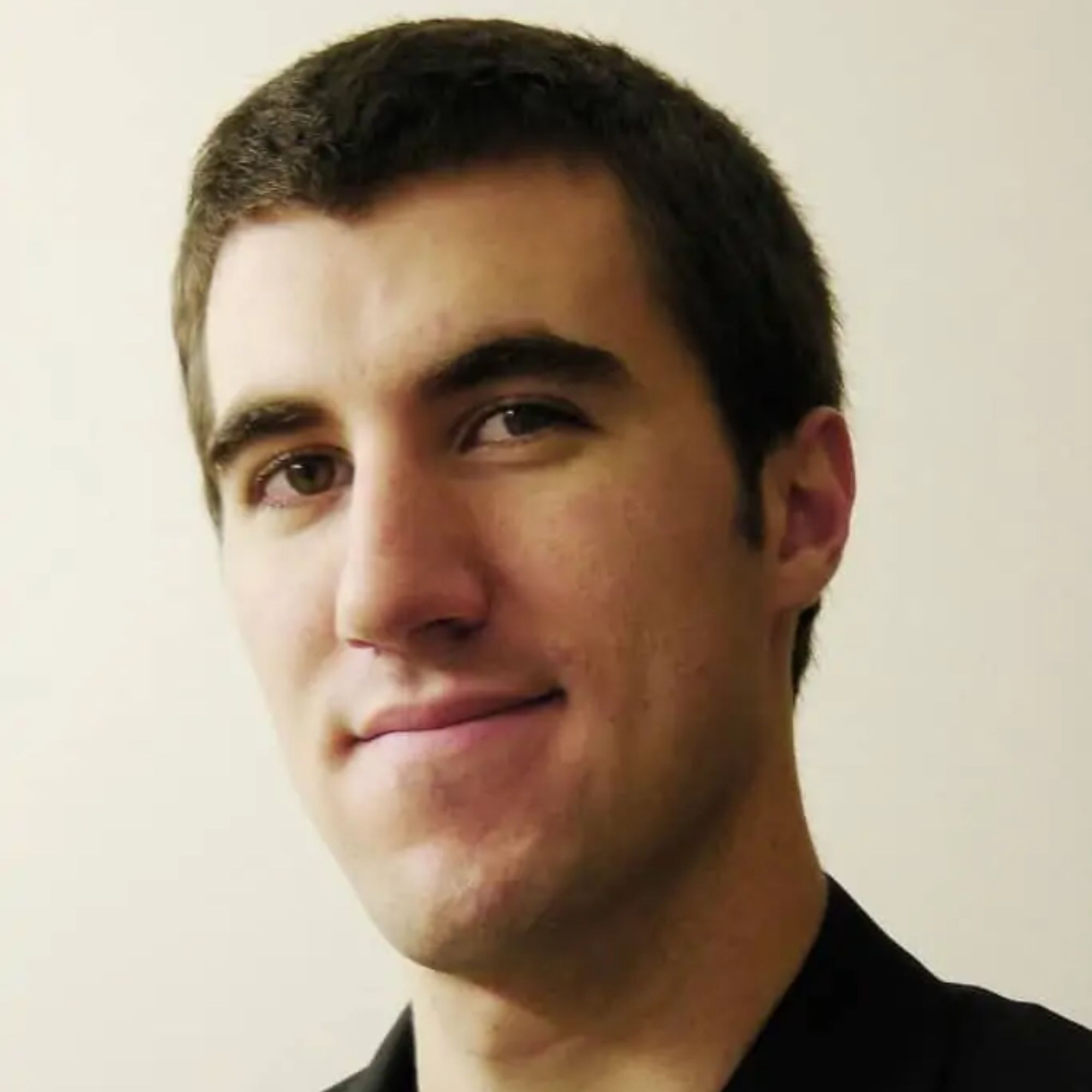 A headshot of a young man with short dark hair wearing a black top, smiling slightly against a cream background