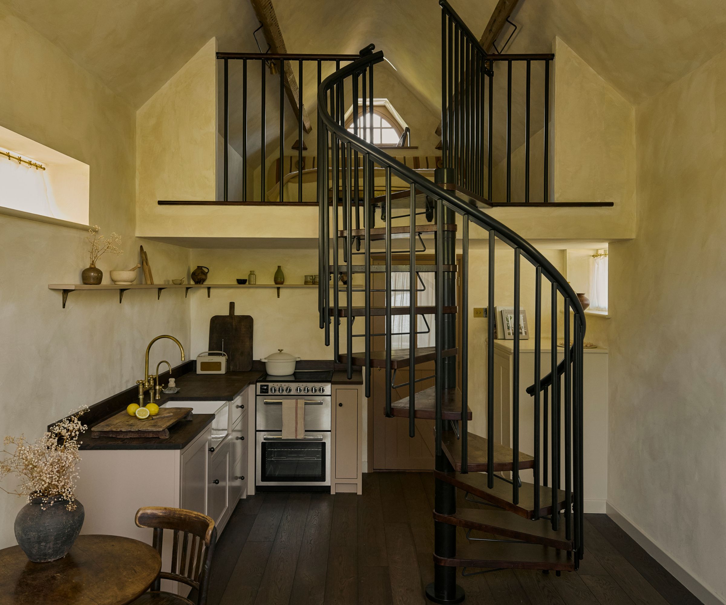 The inside of a tiny home with warm neutral limewash walls, a black spiral staircase, a small L-shaped kitchen with wooden countertops, and a mezzanine floor for the bedroom