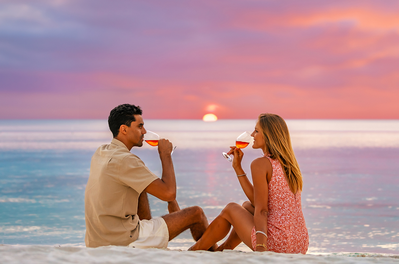 A couple sitting on white sand drinking a glass of ros&amp;eacute; at sunset, the sky is pink