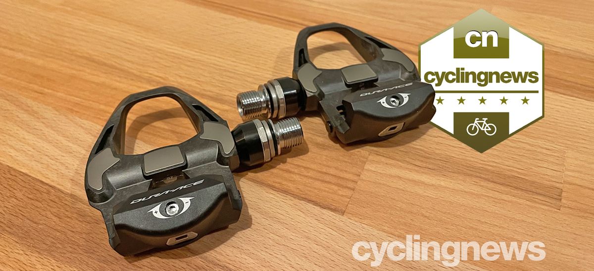 Two Shimano Dura-Ace pedals on a wooden surface, overlaid with a five star badge