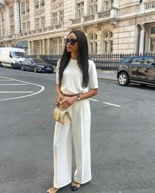 Woman wearing white tailored trousers, white t-shirt, nude ballet flats and black sunglasses while carrying neutral mini bag.