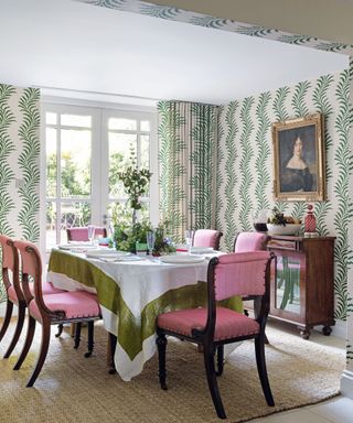 Dining room with green botanical wallpaper, matching curtains, pink dining chairs and natural rug