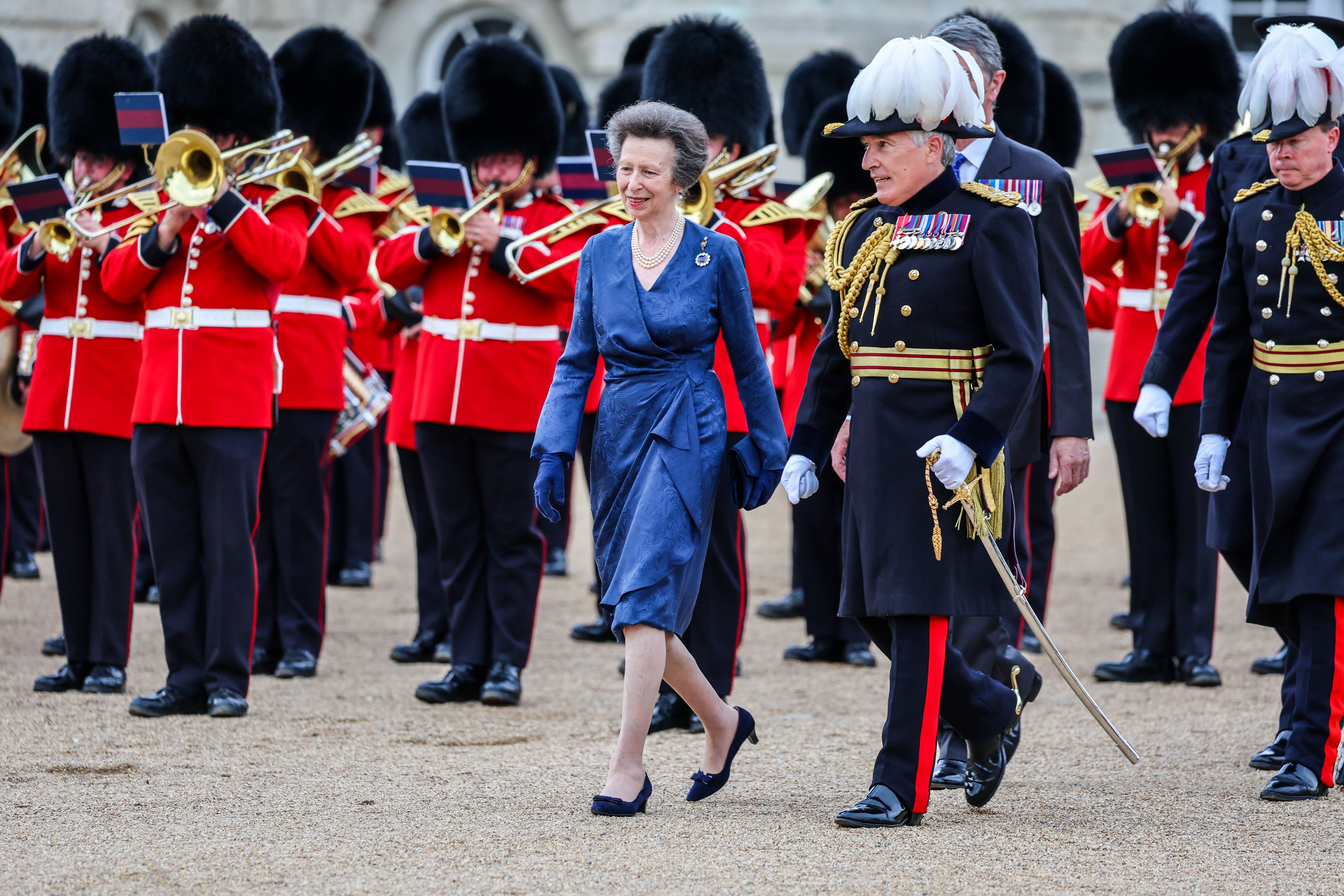Princess Anne wearing a blue dress and heels walking with soldiers and a band