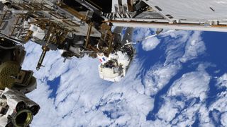 a person in a bulky white spacesuit clings to metal bars attached to a large structure, while white clouds in a blue sky can be seen far below