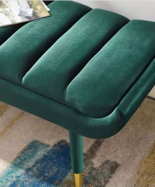 A dark green velvet bench with gold feet on a carpet