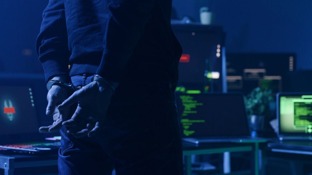 A man in handcuffs standing in front of computer equipment in a darkened room