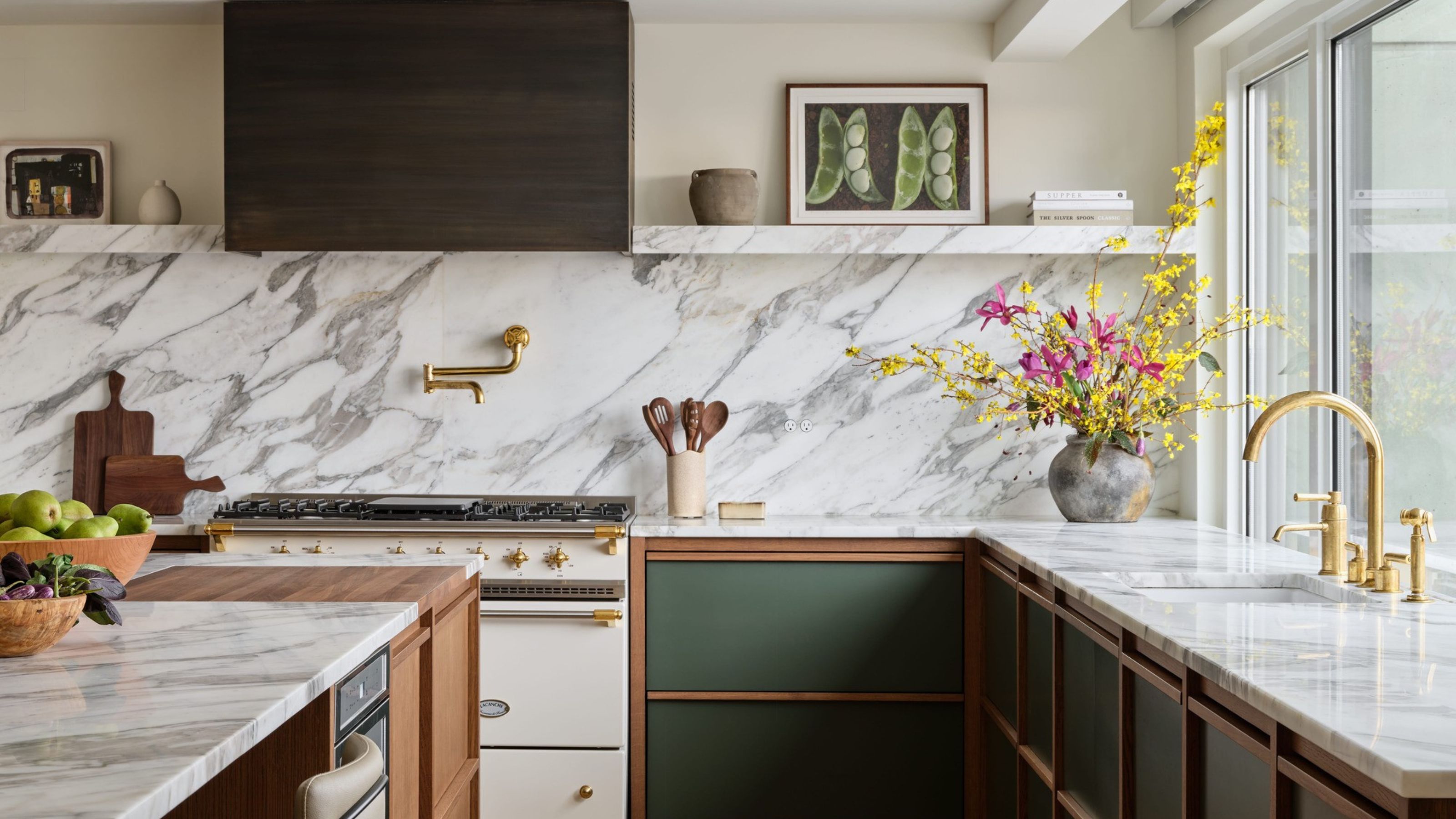 modern green kitchen with marble countertop and backsplash, aga stove, timber range hood, gold taps, vase of flowers, artwork on open shelves, and fruit bowl