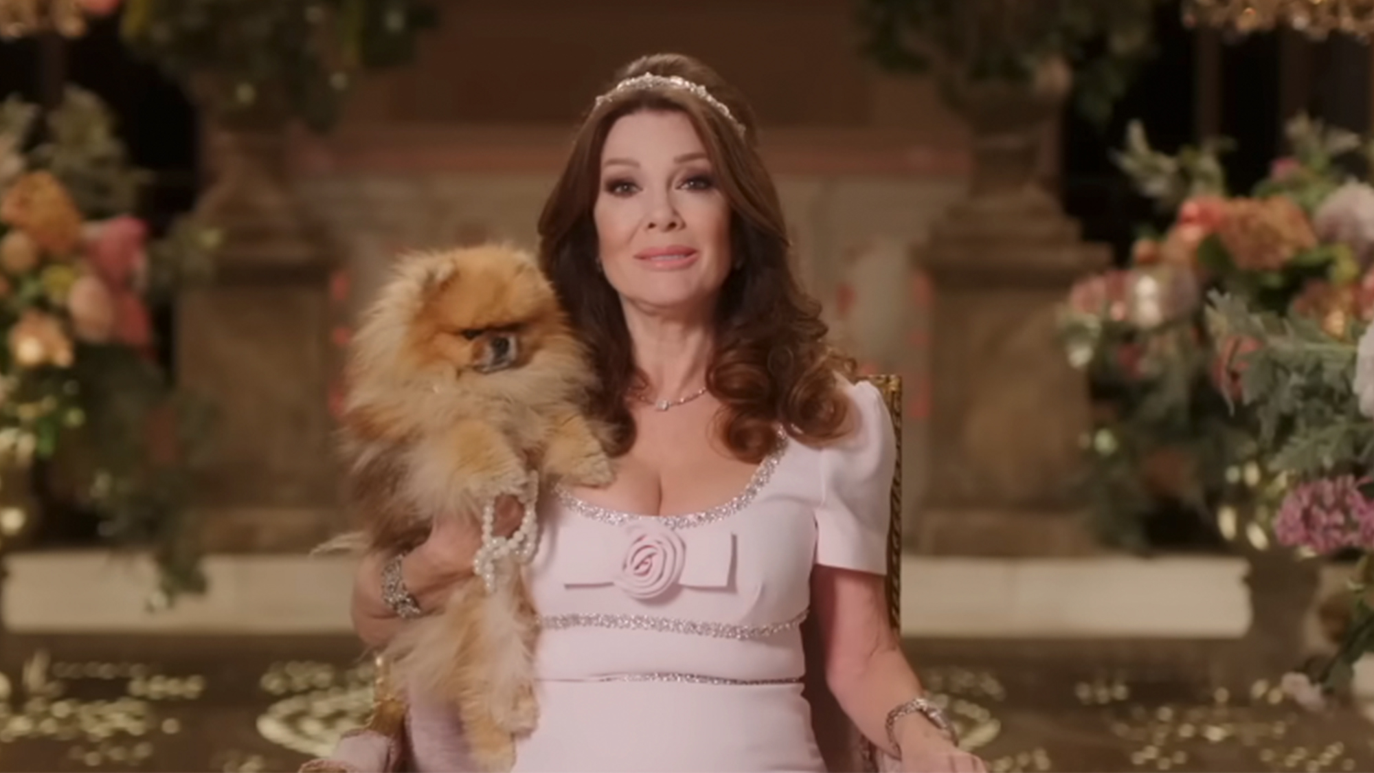 Lisa Vanderpump in a light pink dress and a tiara, holding her dog.
