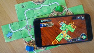 The board game Carcassonne lays on a table in the background with the Carcassonne mobile app displayed in front. 