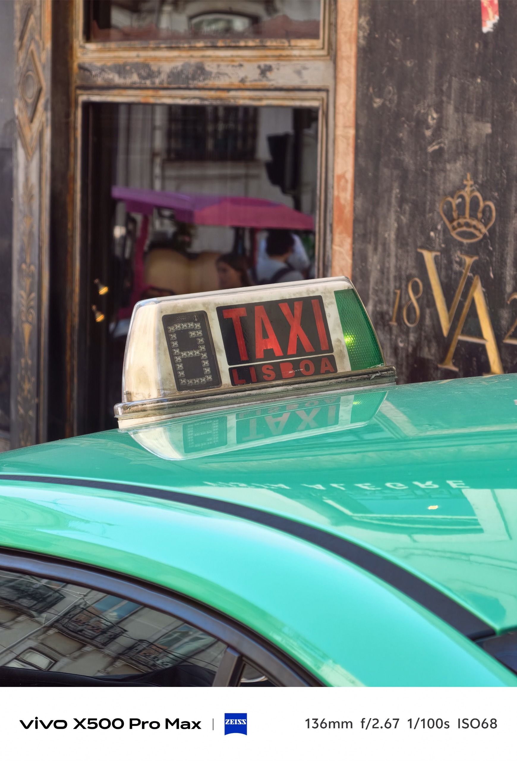 Close view of the roof sign on a turquoise Lisbon taxi, with an ornate building facade behind.