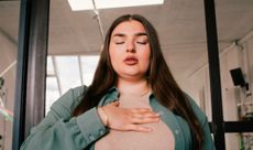 Woman doing breathing exercise. She stands in front of a glass door with her eyes closed, lips slightly open and hand resting on her chest. 