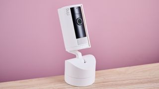 The Ring Pan-Tilt Indoor Cam sits on light-colored wood surface against a pink background.