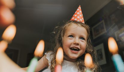 A young blonde girl wearing a birthday hat and smiling as she prepares to blow out some candles during a lockdown birthday for kids.