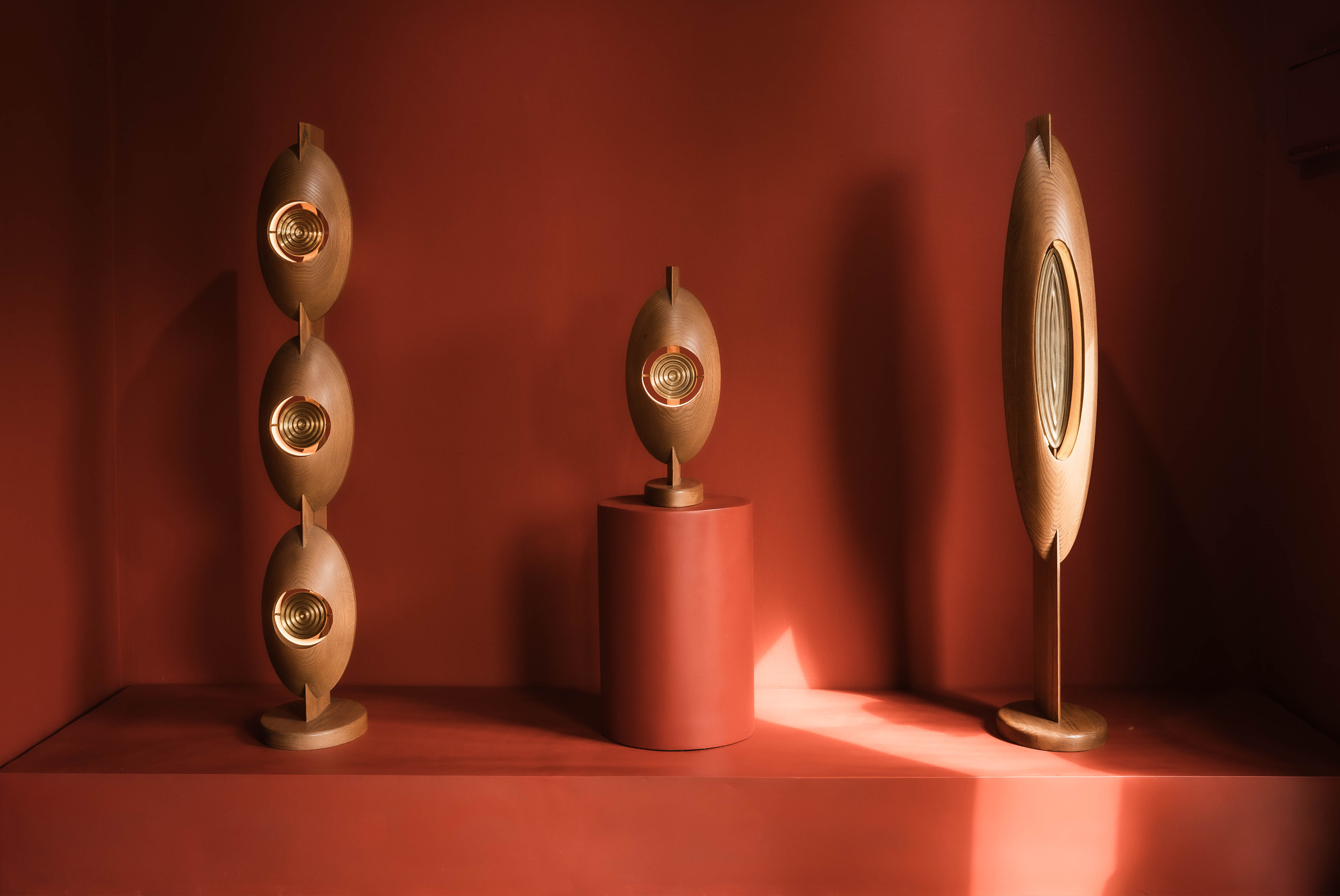 A red-painted gallery room features a trio of wooden lights sculpted in drop-shaped silhouettes with golden inserts atop podiums.