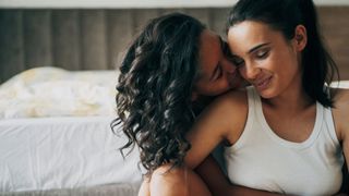 Two women hugging each other, sitting at the foot of their bed