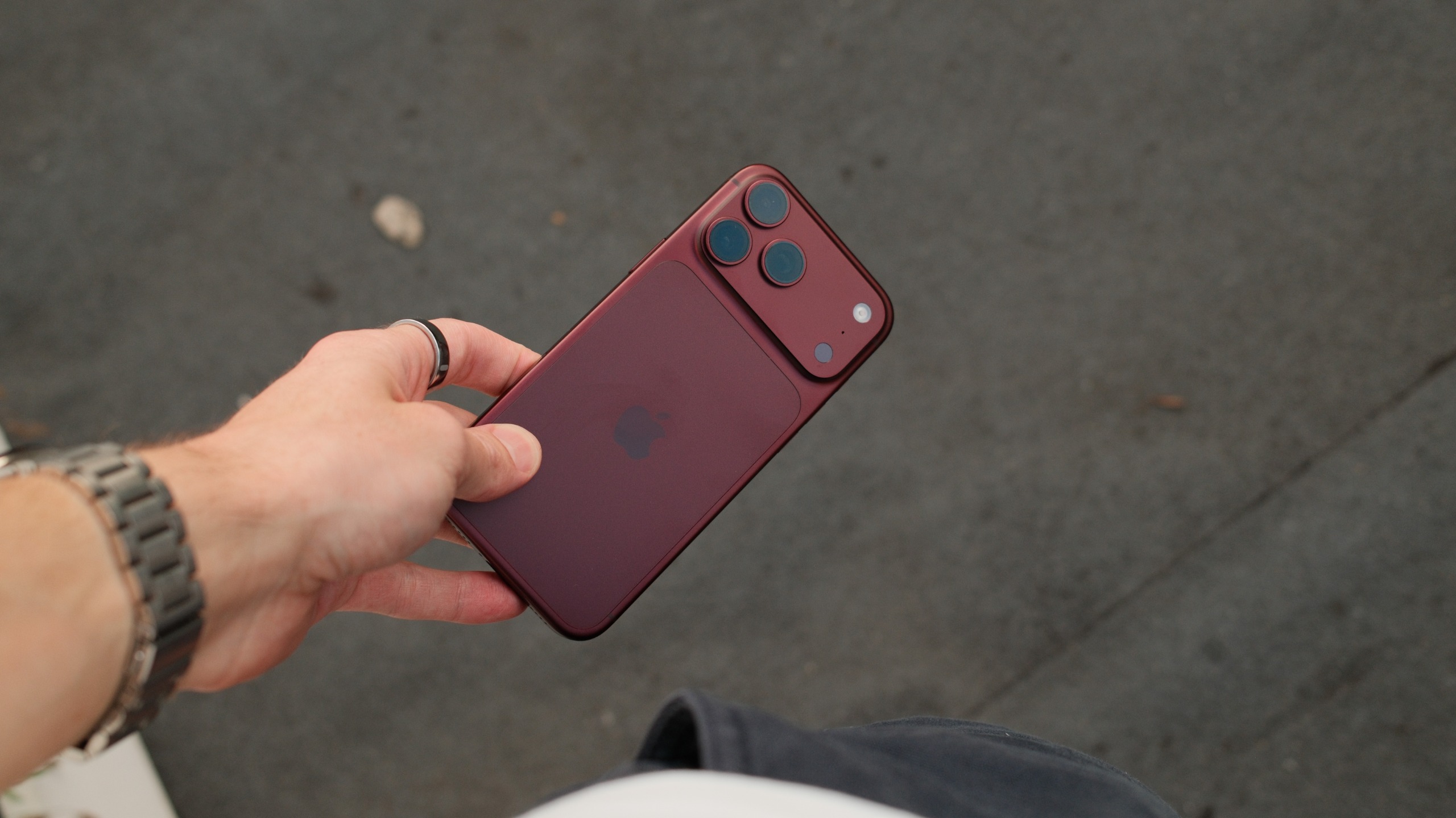 Burgundy iPhone 18 Pro Max held over a pavement, showing the matte rear finish, Apple logo, and triple cameras.