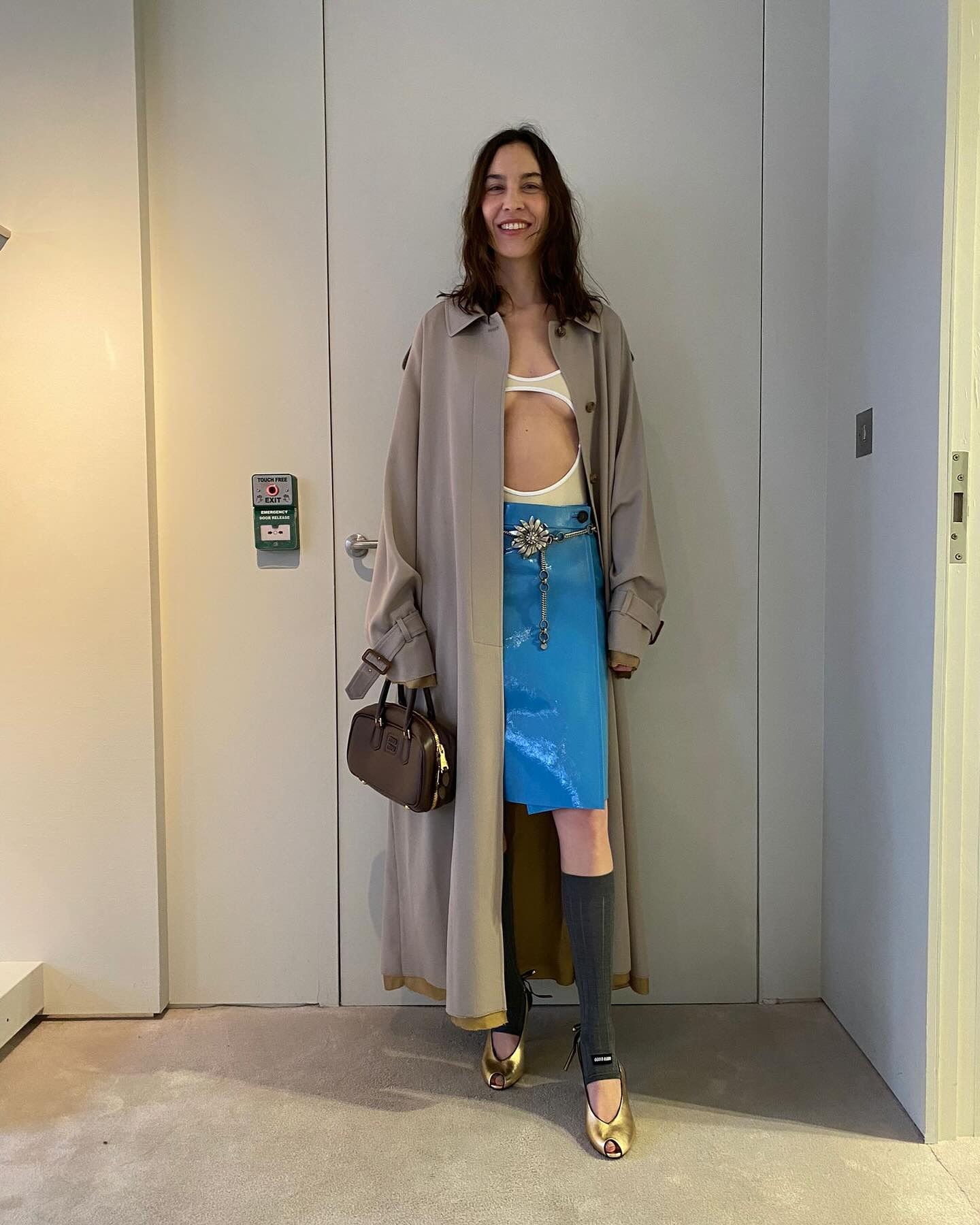 Alexa Chung wearing a trench coat with a swimsuit and patent leather blue Miu Miu skirt with knee-high suit socks and gold peep-toe heels.