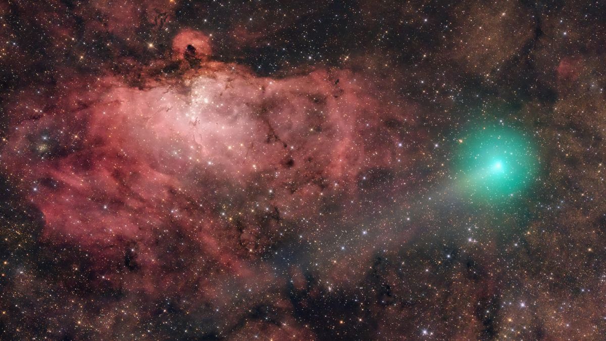 Se puede ver un cometa verde brillando a la derecha de una imagen del espacio profundo, con una cola tenue que se extiende hacia la parte inferior izquierda. Se puede ver una nebulosa roja arremolinada dominando el fondo a la izquierda de la pantalla, con una serie de estrellas particularmente brillantes agrupadas debajo de un denso filamento horizontal de polvo cósmico.