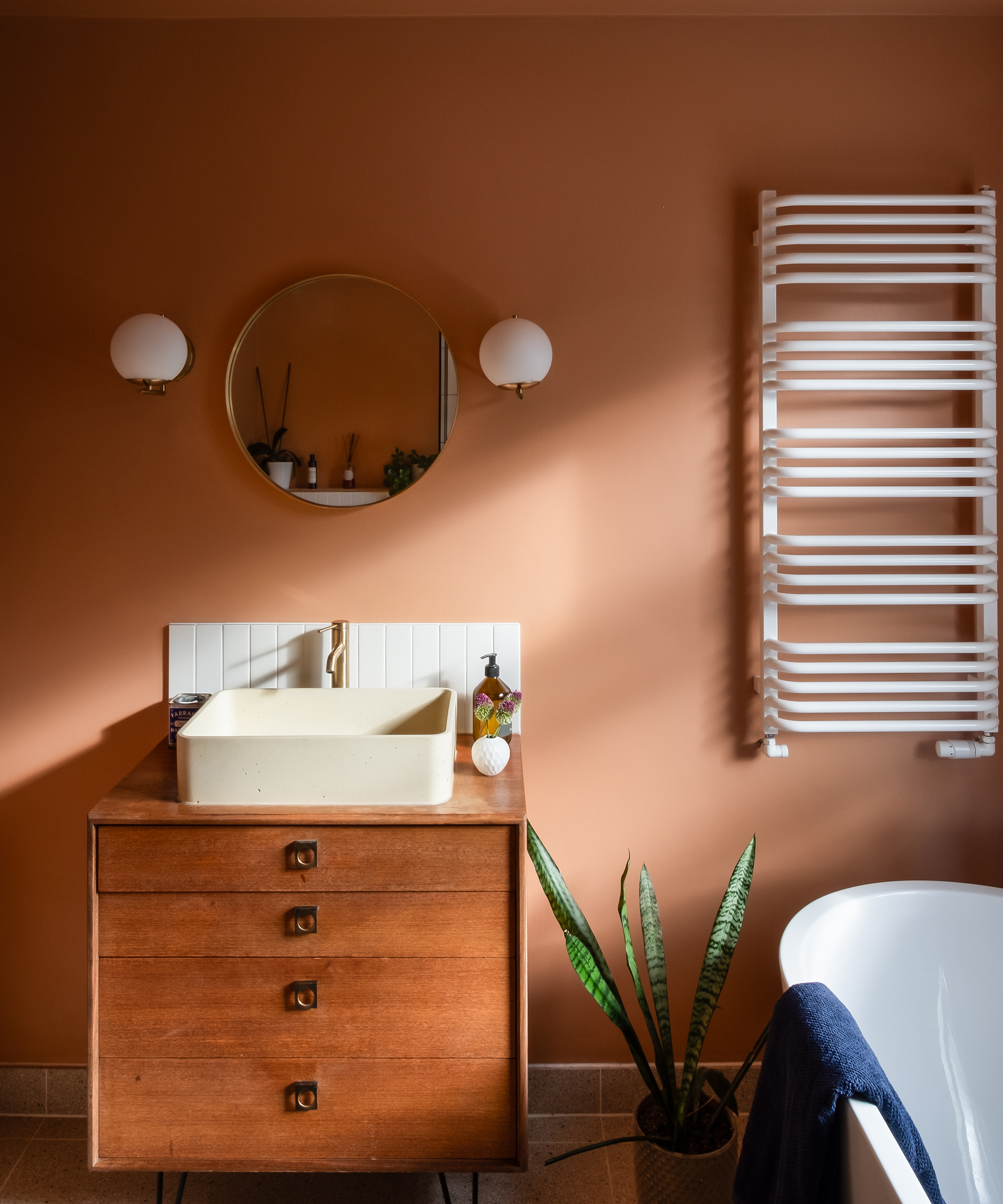 A peach painted bathroom with a wooden set of drawers as the sink unit. There is a white towel rail on the wall next to it and a roll top bath to the right of the image
