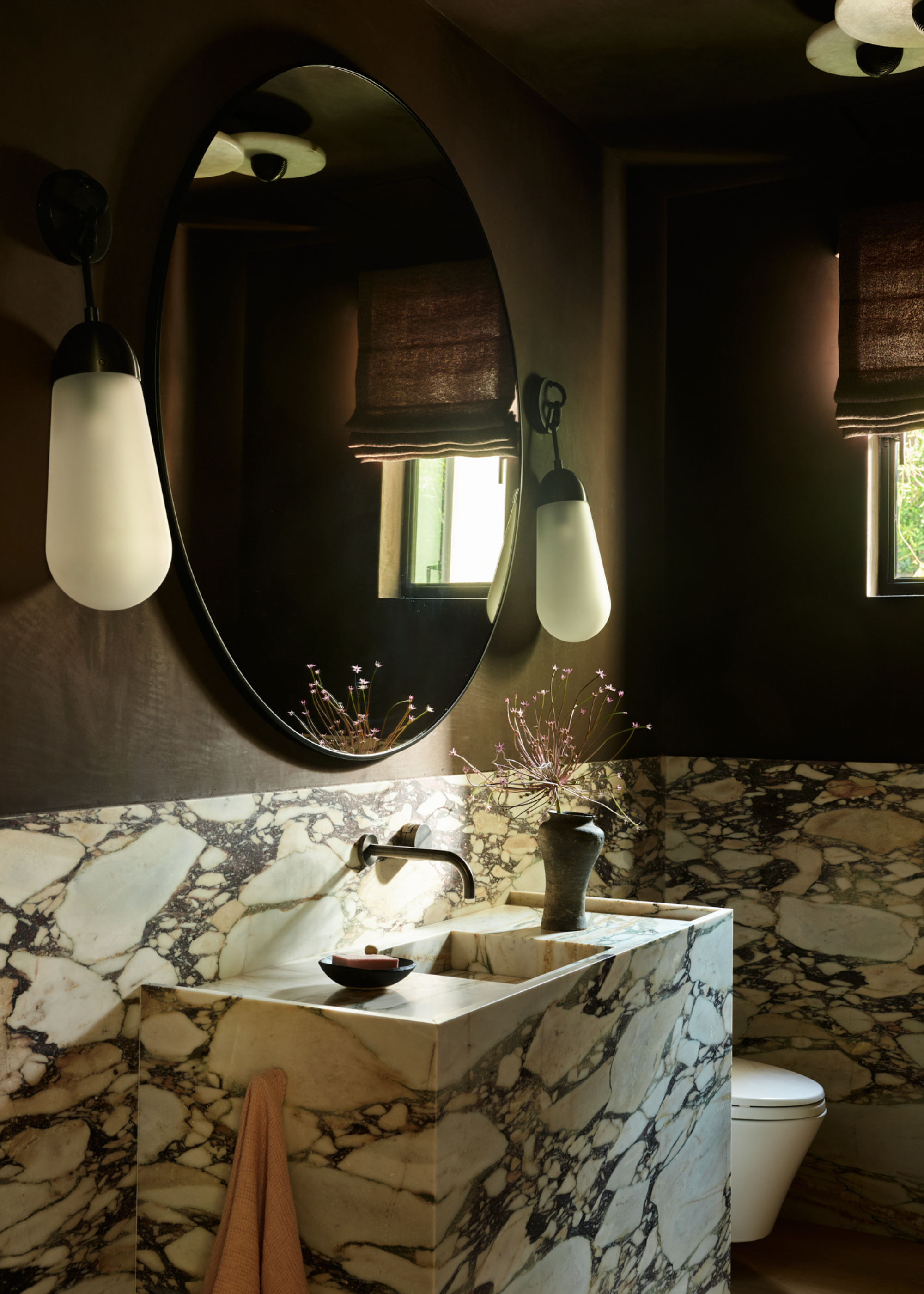 A powder room with dark brown walls, a round mirror, hanging sconces, a marble sink and a toilet