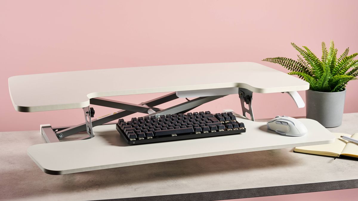 The Flexispot M17 sits on a stone-effect desk in front of a pink background. There&#039;s a keyboard and mouse on the desk, and a house plant and notebook to the right.