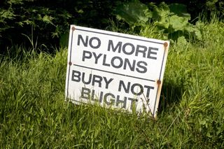 A sign on a grass roadside verge reading 'No More Pylons - Bury Not Blight' protesting against pylons