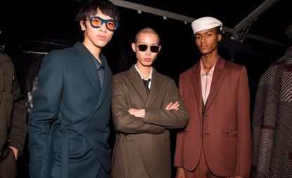 Three male models stand shoulder to shoulder wearing various suit jackets and accessories