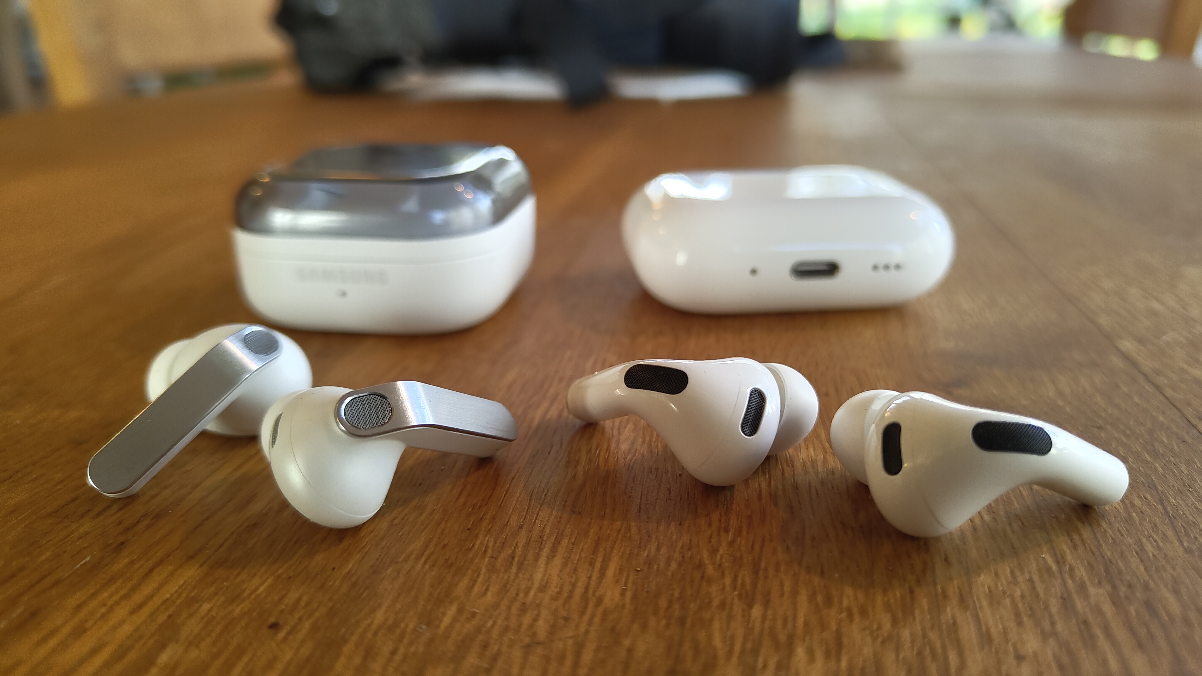 Samsung Galaxy Buds 4 Pro vs Apple AirPods Pro 3 side by side on a wooden table