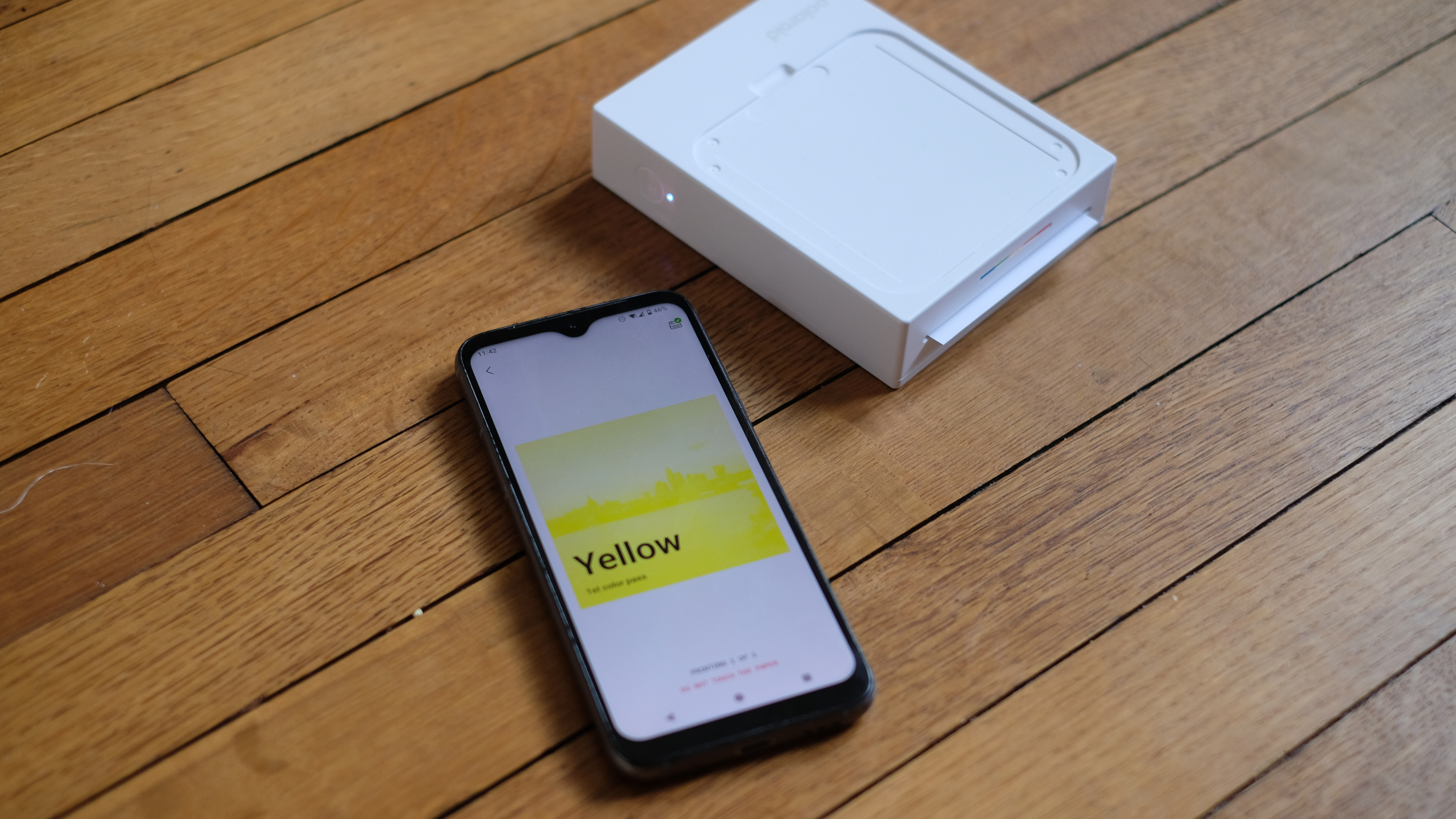 Photograph of Polaroid Hi-Print 3x3 mini instant printer mid-print, with app connected visible on smartphone screen.