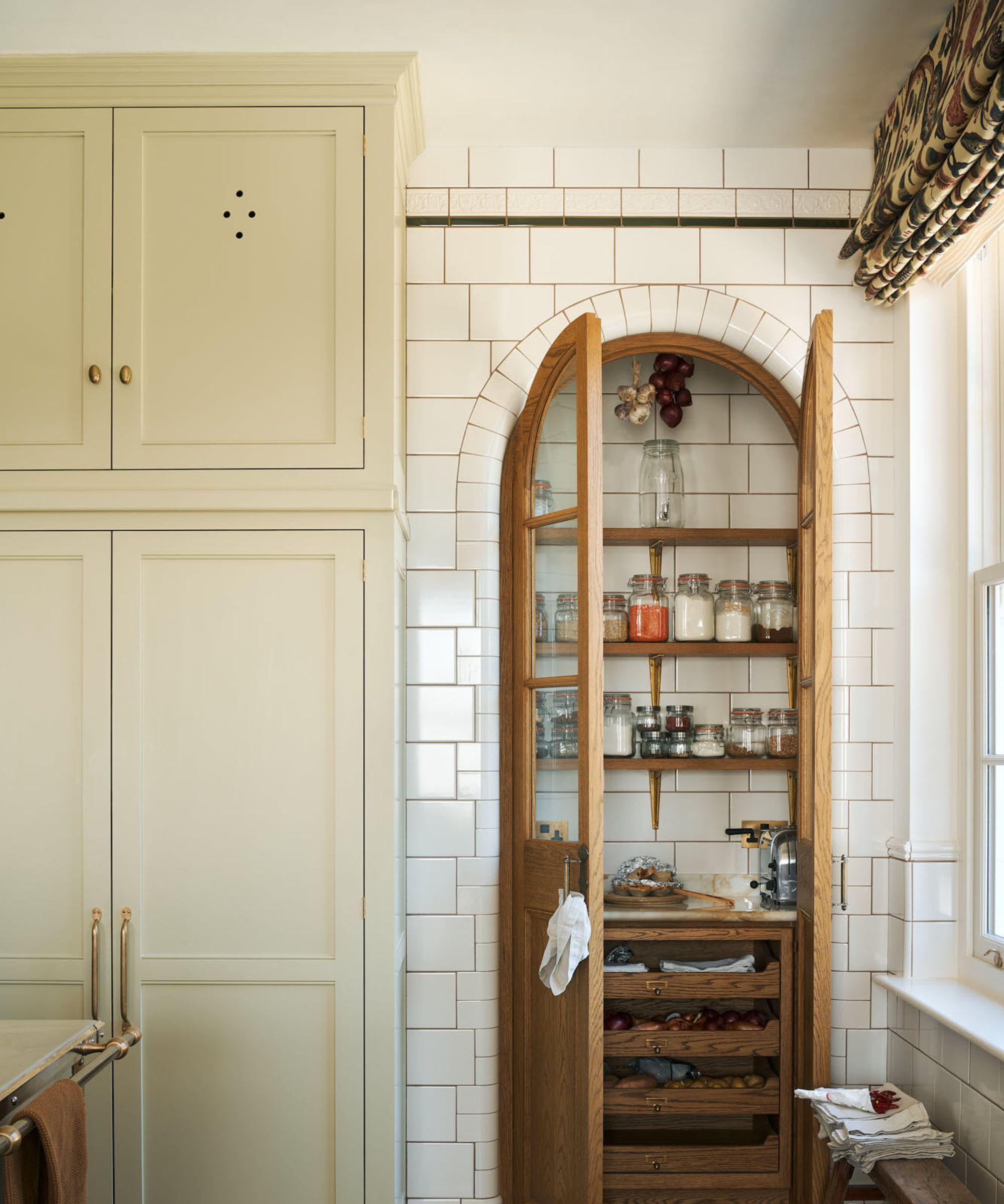 The corner of a neutral kitchen with an arched wood pantry door next to cream cabinets with cutout details