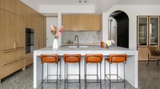 Image of a white kitchen with wooden cabinetry and a large white island. There are four orange and metal bar stools. There is a white vase with pink flowers on the counter.