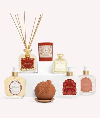 A series of gold and red perfume bottles, diffusers, candles, soaps appear alongside a ceramic pomegranate against a white background.