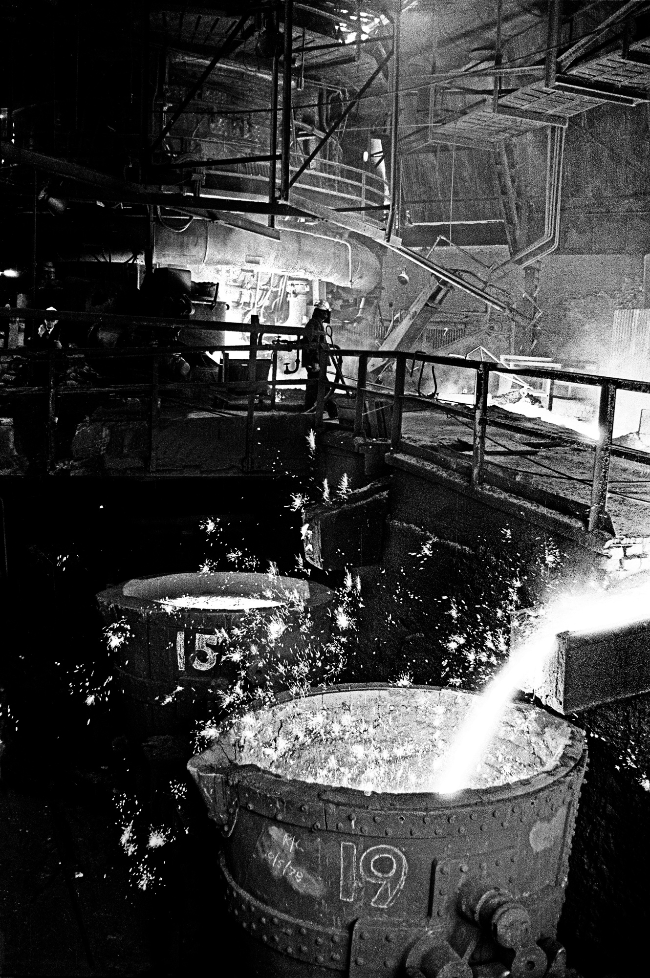Black and white photograph of a steelworker in protective gear silhouetted beside a furnace as molten metal glows and sparks fly in the dark