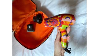 Original image showing hand-held close-up of the Amika Mighty Mini Dryer, its attachments and case