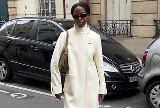 French influencer wearing white funnel-neck coat with brown back and black sunglasses, standing on Paris street.