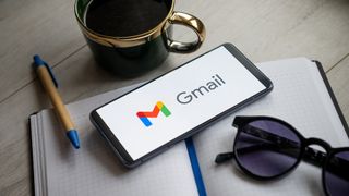 Gmail branding and logo pictured on a smartphone sitting on a desk alongside coffee cup and notebook.