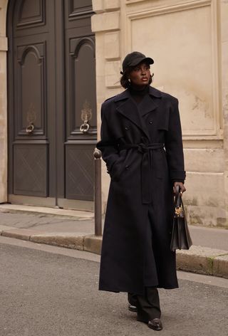 best long coats are shown in a photo of a long coat outfit with a woman standing on the sidewalk wearing a black baseball cap with a black turtleneck, navy trench coat, black trousers, black loafers, and a black bag