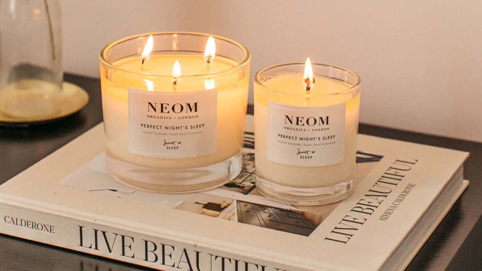 Two NEOM Perfect Night's Sleep candles burning on a white coffee table book on a dark wood table against a white wall